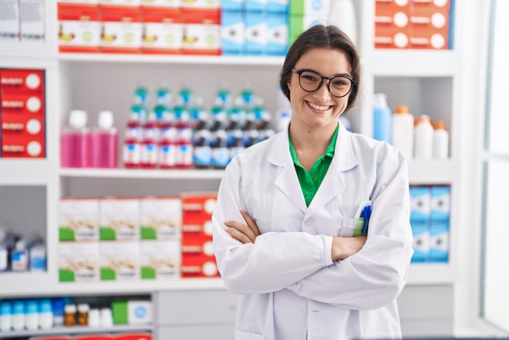 Jovem farmacêutica hispânica sorrindo confiante em pé com os braços cruzados em gesto na farmácia